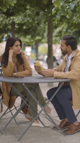 Video-Vertical-De-Una-Pareja-Musulmana-En-Una-Cita-Sentada-En-Una-Mesa-Al-Aire-Libre-En-Una-Calle-De-La-Ciudad-Intercambiando-Números-De-Teléfono-1